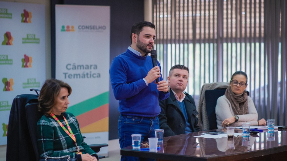 Gabriel em pé, atrás de uma mesa falando ao microfone e sendo olhado por mais três pessoas que compõem a mesa junto com ele.